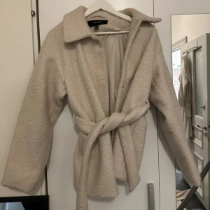 Beige teddyjacka med bälte från Vero Moda - Mysig beige teddyjacka från Vero Moda med bred krage och knytskärp i midjan. Jackan har lång ärm och en oversized look, perfekt för att hålla värmen under kyliga dagar. Materialet är fluffigt och mjukt, vilket ger en trendig och avslappnad vibe.