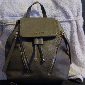 Oliv ryggsäck från G by Guess - Snygg grå ryggsäck från G by Guess i strukturerat skinnimitation. Väskan har två dragkedjefickor framtill, snörning upptill och justerbara axelremmar. Perfekt storlek för vardagens alla prylar och stilren design med metallogo på locket.