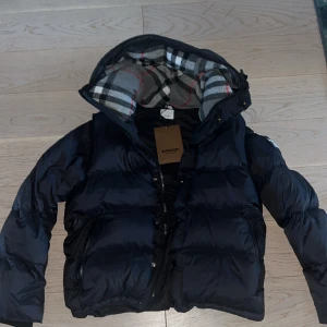 Svart pufferjacka från Burberry - Pufferjacka från Burberry med avtagbara ärmar. Jackan har dragkedja som går upp till näsan, knappar och stor logotyp på ena ärmen. Perfekt för kalla dagar och kan bäras som väst. Säljer eftersom jag redan har en annan vinterjacka, pris kan diskuteras.
