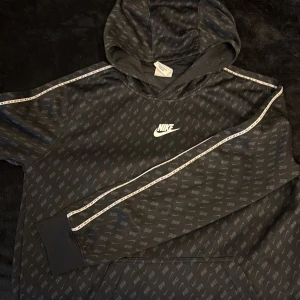 Svart hoodie från Nike med logomönster - Svart hoodie från Nike med all-over logomönster och vit Nike-logga på bröstet. Tröjan har huva, långa ärmar och vita detaljer längs axlar och ärmar. Tillverkad i mjuk polyester, perfekt för chill eller träning.