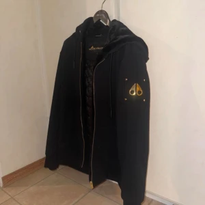 Svart pufferjacka från Moose Knuckles - Svart pufferjacka från Moose Knuckles med guldfärgade metalldetaljer på ärmen och dragkedja. Jackan har huva, quiltat innerfoder och snygga detaljer som ger en exklusiv känsla. Perfekt för dig som vill ha en varm och stilren jacka med streetkänsla.