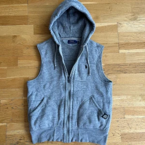 Grå fleeceväst från Polo Ralph Lauren - Snygg grå väst med huva från Polo Ralph Lauren. Västen har dragkedja framtill, två fickor och justerbara snören i huvan. Tillverkad i bomull och polyester med ribbade detaljer vid axlar och huva. 