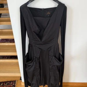Svart långärmad klänning Vivienne Westwood - Svart långärmad klänning från Vivienne Westwood med markerad midja och stora fickor på sidorna. Klänningen har dragkedja bak och är tillverkad i 100% bomull med en snygg siluett. Använd fåtal gånger. OBS! Motsvarar en svensk 36 i storlek.