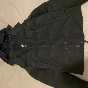 Svart pufferjacka från Canada Goose - Svart pufferjacka från Canada Goose med huva och klassisk logga på insidan. Jackan har dragkedja och tryckknappar framtill, samt flera praktiska fickor. Perfekt för kalla vinterdagar och har en snygg, stilren look.