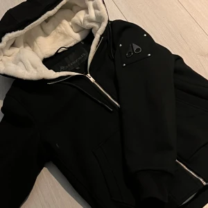Svart Moose Knuckles Bunny-Zip - Svart zip från Moose Knuckles med vit fluffig huva och quiltat innerfoder. Jackan har dragkedja framtill, ribbade muddar och en snygg logotyp på ärmen. Perfekt för kalla dagar och har en riktigt skön vibe. Använd 2 gånger 