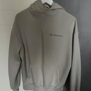 Beige hoodie från Bohoo - Snygg beige hoodie  med minimalistiskt tryck på bröstet. Tröjan har en stor huva, magficka och ribbade muddar vid ärmslut och nederkant. Perfekt för chill dagar och enkel att matcha med jeans eller joggers.