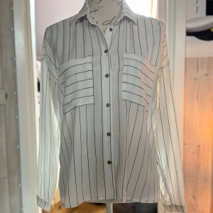 Vit randig skjorta från Rut&Circle - Säljer en vit skjorta från Rut&Circle i storlek 36 med svarta tunna ränder. Skjortan har två bröstfickor, knappar framtill och klassisk krage. Långärmad och i ett lätt, luftigt material som känns skönt mot huden.
