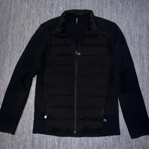 Sail Racing Cardigan - Svart jacka med quiltad, lätt vadderad front och släta ärmar. Jackan har dragkedja hela vägen och hög krage. Materialmix med vadderad polyester framtill och stickade partier på ärmar och rygg. Perfekt för lager-på-lager och chill stil.