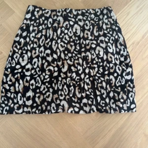 Leopardmönstrad kortkjol från H&M - Snygg kortkjol från H&M med leopardmönster i svart, beige och brunt. Kjolen har en liten slits framtill och är fodrad på insidan. Perfekt för dig som vill sticka ut med ett djurmönstrat plagg.