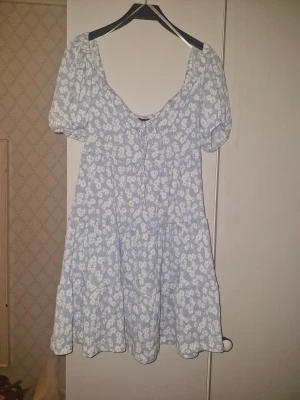 Blommig ljusblå klänning Gina Tricot XS - Supersöt ljusblå kortklänning från Gina Tricot i storlek XS. Klänningen har puffiga kortärmar, v-ringning med knyt framtill och ett vitt blommigt mönster. Tillverkad i mjuk bomull, perfekt för sommarens alla tillfällen. Volangdetaljer ger en extra feminin touch.