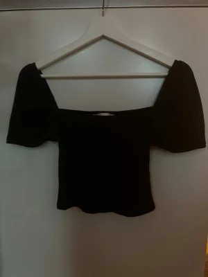 Svart topp - Säljer en svart topp med korta puffärmar. Toppen är croppad och har en stilren, enkel design som passar perfekt till jeans eller kjol. Materialet känns mjukt och stretchigt, vilket gör den bekväm att bära. Perfekt för dig som gillar trendiga och snygga plagg.