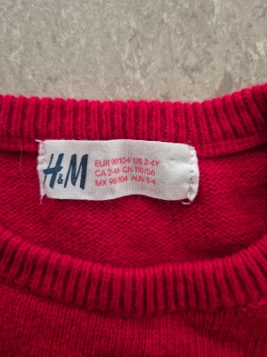 Stickad tröja - Snygg röd stickad tröja från H&M i storlek 98/104. Tröjan har ett stort hjärta framtill gjort av glittriga paljetter och puffade axlar som ger en cool look. Rund hals och långa ärmar med ribbade muddar. Perfekt för dig som gillar färg och glitter!