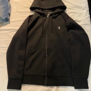 Svart hoodie från Polo Ralph Lauren - Svart hoodie från Polo Ralph Lauren med dragkedja framtill, snörning vid huvan och klassisk logga broderad på bröstet. Tröjan har två fickor och är tillverkad i mjuk bomullsmix. Perfekt för chill dagar eller när du vill ha en clean look.