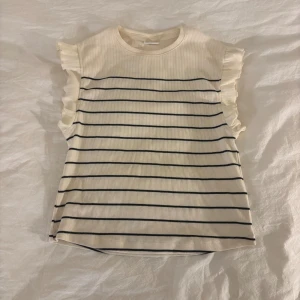 Randig vit blus med volang från Zara - Supersöt blus från Zara i ribbad bomull, vit med tunna svarta ränder och volangdetaljer på ärmarna. Blusen är ärmlös med rund hals och har en mjuk, lätt stretchig känsla💕