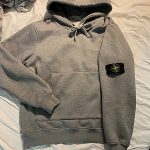 Grå hoodie från Stone Island - Snygg grå hoodie från Stone Island med klassisk logotyp-patch på vänster ärm. Tröjan har huva med snörning och en stor magficka framtill. Perfekt för dig som gillar streetwear och vill ha en clean look.