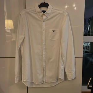 Vit Oxfordskjorta från GANT, L - Vit klassisk Oxfordskjorta från GANT i storlek L, med button-down krage och bröstficka med broderad logga. Skjortan har lång ärm, knappar framtill och är tillverkad i mjuk bomull. Regular fit för en avslappnad och snygg look.