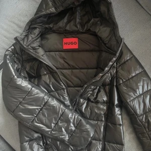 Svart pufferjacka från Hugo Boss - Svart pufferjacka från Hugo Boss med glansig finish och huva. Jackan har quiltad design, dragkedja framtill och två sidofickor med dragkedja. Märket syns tydligt på bröstet och insidan. Perfekt för kalla dagar och har en modern, clean look. Ja kan är i köpt våren 25. Kostade 2000:-