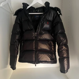 Svart pufferjacka från Polo Ralph Lauren - Snygg svart pufferjacka från Polo Ralph Lauren med glansig finish, huva och dragkedja framtill. Jackan har två stora fickor med dragkedja och ikonisk röd logga på bröstet. Perfekt för kalla dagar när du vill hålla stilen.