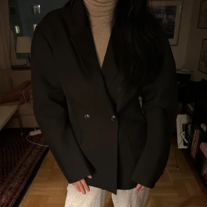 Svart kavaj från Gina Tricot - Stilren svart kavaj från Gina Tricot med klassisk krage och två knappar framtill. Kavajen har raka fickor och en enkel, tidlös design som passar till många olika looks. Perfekt för dig som gillar en clean och snygg stil.