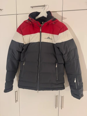 J.Lindeberg dunjacka - Säljer en snygg pufferjacka från J.Lindeberg med breda färgblock i rött, vitt och mörkgrått. Jackan har huva, flera dragkedjor och logga på bröstet och ärmen. Perfekt för dig som vill ha en varm och stilren jacka med sportig vibe.
