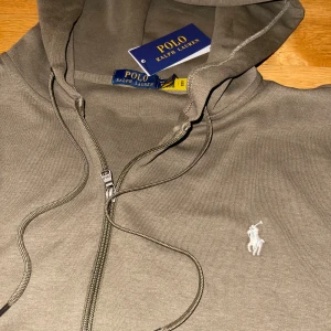 Grå hoodie från Polo Ralph Lauren - Snygg grå hoodie från Polo Ralph Lauren med dragkedja och huva. Tröjan har snörning vid halsen, två fickor fram och den klassiska Polo-loggan broderad på bröstet. Perfekt för chill dagar och enkel att matcha med jeans eller joggers.