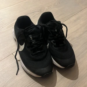 Barnskor Nike  - Säljer ett par Nike sneakers med vit sula och stor vit swoosh på sidan. Skorna har snörning och är tillverkade i mesh och syntetmaterial för bra ventilation och komfort. Sulan är mönstrad för extra grepp och dämpning, perfekt för träning eller promenader.
