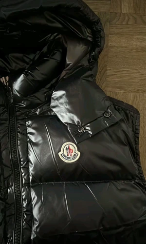 Svart dunväst från Moncler med huva - Säljer en svart, glansig dunväst från Moncler med stor huva och två dragkedjeförsedda fickor. Västen har ett quiltat mönster och Moncler-logga på bröstet. Perfekt för kalla dagar när du vill ha både stil och värme.
