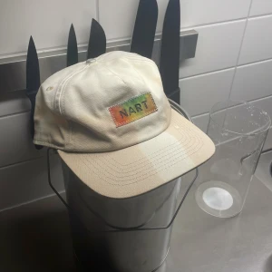 NART Keps - Unik beige keps från NART, designad i Stockholm och färdigställd 2024-10-25. Framtill sitter en färgglad patch med broderad text och zigzag-söm. Kepsen har böjd skärm och justerbar rem bak. Perfekt för dig som gillar limited edition och streetstyle.