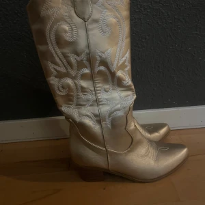 Guldiga cowboy boots med broderi - Snygga cowboy boots i metallic guld med vit broderad dekor längs skaftet och framtill. Skorna har spetsig tå, låg klack och är tillverkade i syntetmaterial med skinnliknande finish. Perfekt för dig som vill sticka ut med snygga boots.