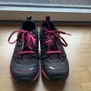 Icebug Pytho3 Bugrip - Säljer ett par  vattentäta svarta Icebug skor med rosa snörning och detaljer. Skorna har grov mönstrad sula med metall-dubbar för extra grepp. Materialet är syntet och textil, med en gul logga på sidan och vadderad insida för komfort.