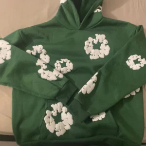 Grön demin tears hoodie - Säljer en grön demin tears hoodie med huva och stora vita blommönster tryckta över hela tröjan. Hoodien har en klassisk passform, lång ärm och en magficka framtill. Perfekt för dig som gillar unika och färgglada plagg med coola detaljer.
