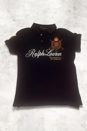 Mörkblå piké från Ralph Lauren - Snygg mörkblå pikétröja från Ralph Lauren Sport med broderad logga och emblem i guld och rött på bröstet. Stora vita bokstäver framtill, klassisk krage och korta ärmar. Materialet är bomull och har två knappar vid halsen. Perfekt för dig som gillar preppy stil.