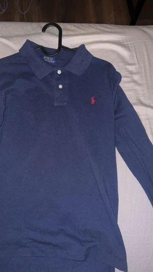Marinblå pikétröja från Ralph Lauren - Snygg marinblå långärmad pikétröja från Ralph Lauren med klassisk krage och tre vita knappar framtill. Tröjan har det ikoniska röda Polo-logot broderat på bröstet och är gjord i mjuk bomull. Perfekt för dig som gillar stilrena och tidlösa plagg.
