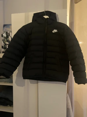Svart pufferjacka från Nike - Svart pufferjacka från Nike med huva och dragkedja framtill. Strl 137-147 cm