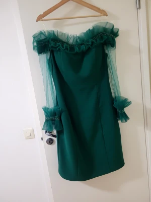 Grön offshoulder klänning med tyll - Säljer en grön kortklänning med offshoulder och transparenta tyllärmar. Klänningen har volangdetaljer och pärlor vid ärmslut och halsringning. Perfekt för dig som vill sticka ut med en unik och elegant look.