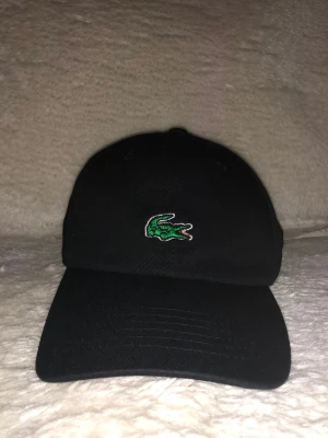 Svart Lacoste keps - Svart keps från Lacoste med klassisk grön krokodillogo broderad framtill och vit Lacoste-text baktill. Justerbar rem i brunt läder med metallspänne. Snygg och stilren design som passar perfekt till streetwear och casual outfits.