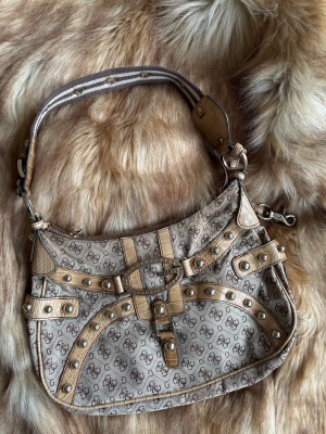 Beige axelväska från Guess med nitar - Snygg beige axelväska från Guess med bruna detaljer och logomönster över hela väskan. Väskan har dekorativa nitar, croco-präglade partier och ett metallspänne framtill. Axelremmen är randig i beige och brunt med metallringar. Perfekt för dig som gillar statement-accessoarer.
