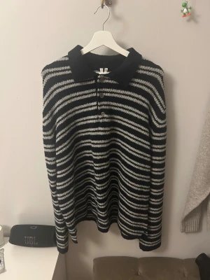 Randig stickad pikétröja i mörkblå och grå/vit från Arket - Säljer en stickad pikétröja i storlek L med mörkblå och gråa ränder. Tröjan har klassisk pikékrage, tre knappar framtill och långa ärmar. Materialet är mjukt och lite fluffigt, perfekt för dig som gillar chill och stilren vibe.