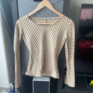 Beige och svart randig långärmad topp Humana - Snygg beige långärmad topp med svarta diagonala ränder. Toppens form är figurnära med rundad hals och mjukt, stretchigt material som känns skönt mot huden. Perfekt för dig som gillar stilrena och trendiga mönster.