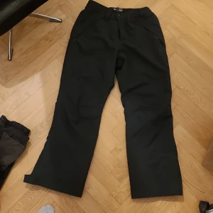 Svarta täckbyxor från Oakley, L/XL - Svarta täckbyxor från Oakley med performance fit, perfekta för vintern. Byxorna har raka ben, knappstängning och dragkedja i midjan samt praktiska fickor med dragkedja. Materialet är slitstarkt syntet, designade för att hålla dig varm och torr.