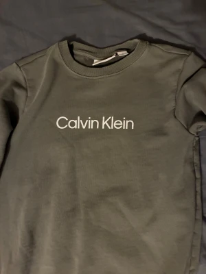 Olivgrön Calvin Klein sweatshirt - Snygg olivgrön sweatshirt från Calvin Klein med vit logotyp broderad på bröstet. Tröjan har rund hals, ribbade muddar och lång ärm. Perfekt för dig som gillar stilrena och klassiska plagg med en modern touch.