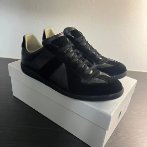 Maison Margiela skor - Maison Margiela sneakers i helsvart med snygga paneler i skinn och mocka. Klassisk rund tå, platt sula och svart snörning. Insidan är ljusbeige och yttersulan har Margielas ikoniska logotyp. Perfekt för dig som gillar minimalistisk och stilren design.