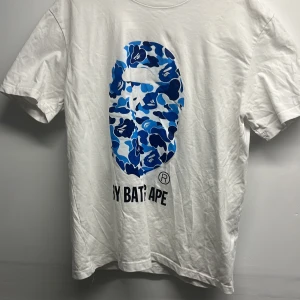 Vit BAPE t-shirt med blå camo print - Säljer en vit t-shirt från BAPE med ikoniskt blått camo-mönster i form av apans ansikte på bröstet. Klassisk rund hals och korta ärmar. T-shirten är gjord i mjuk bomull och har BAPE-logga på ärmen. Perfekt för dig som gillar streetwear och vill sticka ut.