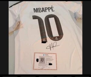 Real Madrid Mbappé fotbollströja M - Säljer en officiell Real Madrid fotbollströja från Adidas i storlek M, med Mbappé och nummer 10 på ryggen. Tröjan är vit med svarta detaljer, gul accent och Real Madrid-emblem. Materialet är lätt polyester med Aeroready-teknologi. Certifikat för äkthet medföljer. Perfekt för fans!
