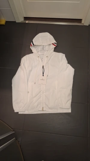 Vit vindjacka från Moncler med huva - Snygg vit vindjacka från Moncler med huva och dragsko. Jackan har Moncler-logga på ärmen, hel dragkedja och elastiska muddar. Insidan av huvan har röd och marinblå rand. Tillverkad i lätt polyester, perfekt för blåsiga dagar.