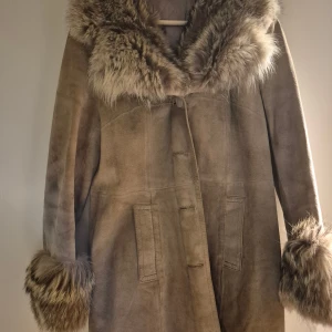Beige mockakappa med fuskpälsdetaljer - Snygg beige vintage kappa i mocka med stora fluffiga fuskpälsdetaljer vid krage och ärmslut. Lång modell med något insvängd midja.Ljust beig ,bilden ej rättvis.