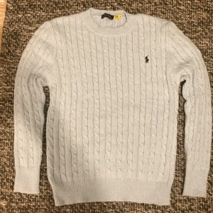 Ljusblå kabelstickad tröja Polo Ralph Lauren - Snygg ljusblå kabelstickad tröja från Polo Ralph Lauren med klassisk rund hals och svart broderad logga på bröstet. Tröjan har långa ärmar och ribbade muddar vid ärmslut och nederkant. 