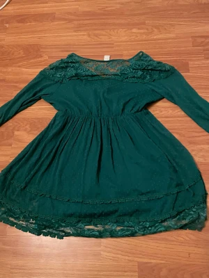 Grön omlottklänning med spets från Vila - Snygg grön kortklänning från Vila med omlottmodell och långärmad design. Klänningen har spetsdetaljer vid axlar och nederkant, samt ett diskret prickigt mönster i tyget. Midjan är markerad och kjolen har volangkant. Perfekt för dig som gillar romantiska detaljer och en feminin look.