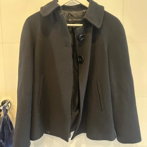 Svart kappa från Zara Basic - Svart kort kappa från Zara. Som man ser, är de två stora knappar vid kragen som går att stänga, den har en fin söm baktill så den lägger sig fint. Den är kort på mig som är 177 cm och speciellt i armarna.