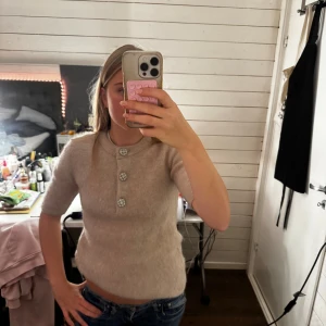 Beige stickad tröja med pärlknappar - Superfin beige stickad tröja från Hm med tre glittriga knappar framtill. Tröjan har korta ärmar och rund halsringning, perfekt för dig som gillar en clean och chic look. Mjuk och skön kvalitet som känns lyxig mot huden.